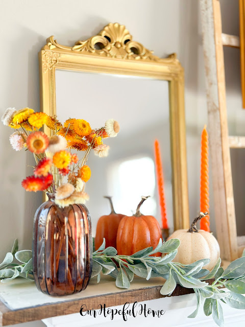amber glass bottle filled with dried orange cone flowers next to orange pumpkin on mantel.