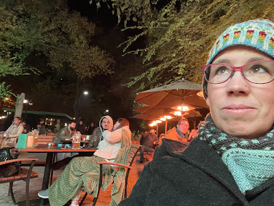 In the foreground, my face is raised to view the screen. I am bundled up in a hat, scarf, and wool coat. The diners visible on the patio behind me are also bundled up.