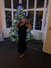 Woman standing in front of Christmas tree wearing a black dress and holding up a sequins mask to her face