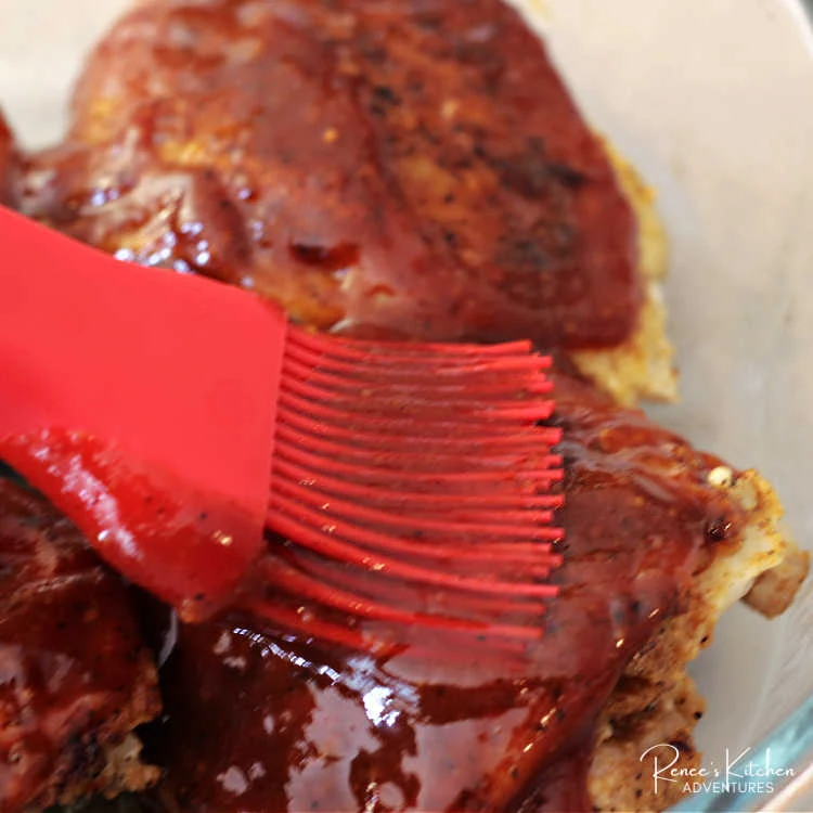 Process photo of chicken thighs in baking dish being basted with homemade sweet and tangy bbq sauce