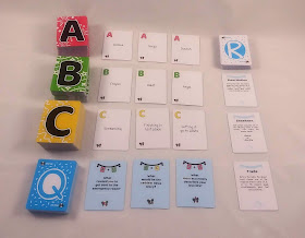 The decks from a game of Cheer Up. Each deck is a different colour with a single large letter that takes up most of the back of the card: A is red, B is green, C is yellow, the question cards have a large Q and are blue, and the rule cards have a large R and are white. Three cards from each deck are laid out next to the decks as examples.