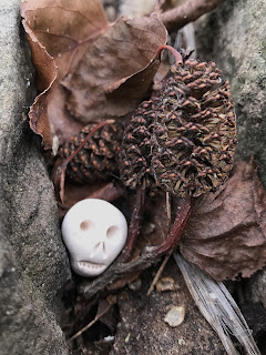 A photo of a small, ceramic skull (Skulferatu #93) lying in a gap in a wall, there are some dead leaves and twigs in the gap along with the Skulferatu.  Photograph by Kevin Nosferatu for the Skulferatu Project.