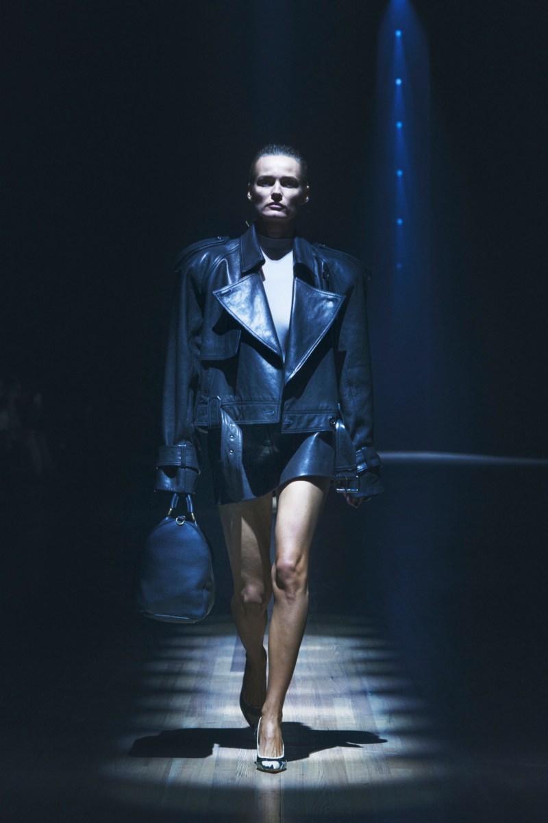 Woman with light brown hair in sleek bun wearing black leather motorcycle jacket, black leather mini skirt, python snakeprint pumps carrying black leather satchel bag walking on a runway with wooden floorboards under a spotlight