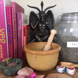Baphomet statue behind a brown mortar and pestle surrounded by pink books, jars of herbs, pink crystals and candles