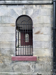 A view through an arched window in the guardhouse.  The window is barred and shows a view through to another barred window and a stone wall in the background.  Photo by Kevin Nosferatu for the Skulferatu Project.