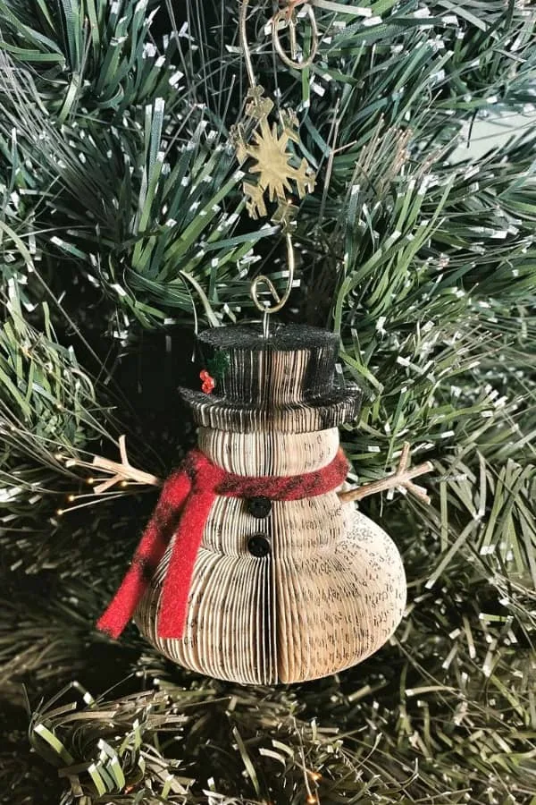 alltered book ornament with twig arms and red and black wooly scarf, hanging on tree