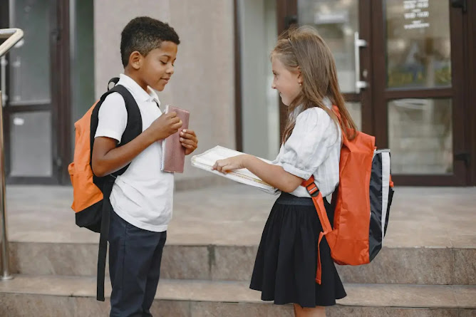 Two schoolchildren, a boy and a girl, standing on stairs outside a building, both wearing backpacks and holding books while engaged in conversation.