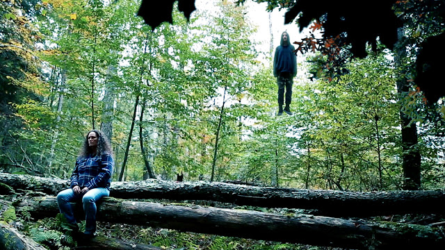 a woman sits on a log while her dead daughter hovers in the background