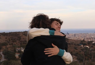Dos jovenes se abrazan fraternalmente en la cima de una colina, y al fondo se observa un valle con una ciudad