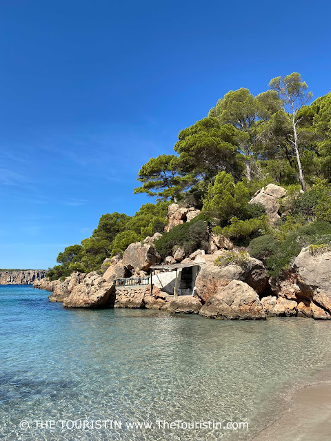 A tiny white cottage on top of a small sandstone wharf surrounded by calm turquoise water and lush greenery under a blue sky.