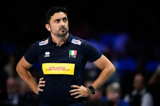 De uniforme azul marinho, Mazzanti está parado com as mãos na cintura e olhando para cima. Ele é moreno, tem cabelo enrolado, barba rala e está aparenta estar irritado