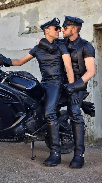 Two Leathermen sitting on motorcycle wearing leather shirts, pants and hats embrace