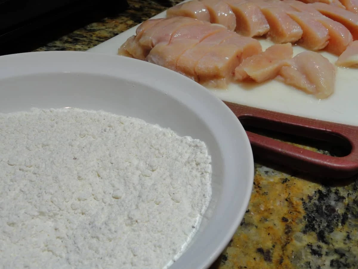 A side shot of a white bowl with seasoned flour and panko bread crumbs with sliced chicken breast in the background.