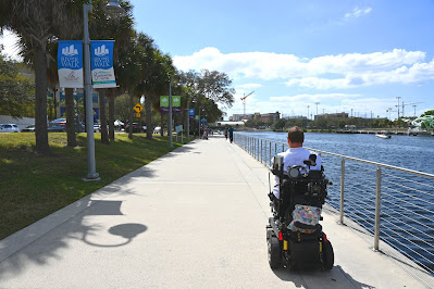 Kyle drives his wheelchair on the right side of a wide sidewalk. To his right is a aluminum guardrail and, on the other side of that, is the Hillsborough River. the sidewalk extends into the distance, where some people can been seen walking on it. To the left is a long patch of grass and a long series of palm trees. A blue sign for Riverwalk hangs on a lamp post, to Kyle's left.