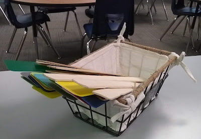 Wire basket, lined with off-white fabric, sitting on library counter-top. In the basket are several 'shelf-markers,' long plastic strips in bright colors and long wooden sticks that are undecorated.