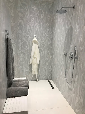 Grey walls of a large shower. On the farthest wall hangs a white, bathrobe. One the wall opposite of the showerhead, lies a bench with two, grey towels resting on it, and a grey towel hanging over it.