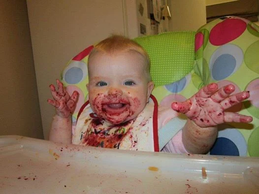 A baby reaching out towards the camera with food on her hands and face and bib from baby led weaning