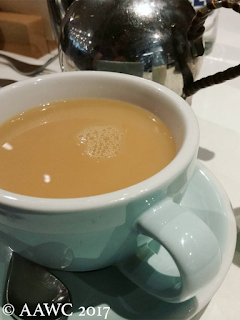 An aqua cup and saucer filled with tea, dusted by milk. A small silver teapot is just visible behind it.