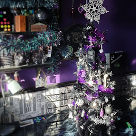 A photograph of Christmas decorations. The wall behind is purple, there is a black dado rail, and greyscale wallpaper of Gothic arches. There are three shelves on the right holding art materials, and the upper two shelves are garlanded with tinsel, grey at the top, iridescent green and purple in the middle. There is a 2 and a half foot tall black Christmas tree densely decorated, with a glittery snowflake on the top that is too large and leans slightly to the right. The tree is on a desk with a fake black marble work-surface. The bottom of the tree is wrapped in a Hallowe'en table-cloth of black lace. The tree is decorated in a purple and silver colour-scheme, with skulls and stars as the primary motifs; it has lots of tinsel. Behind the tree is an ornate black metal stand for a tablet computer.  There is a grey vintage-style desk-lamp on the desk.