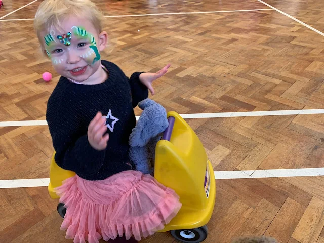 A toddler in party dress, with make up on sitting on a yellow ride on toy and with a blue jelly cat elephant tucked in the seat