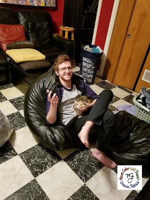 A smiling teenage boy holds a video game controller in one hand and is petting a calico cat under her chin with the other.