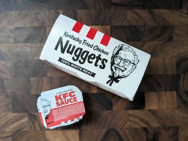 A box of KFC Nuggets next to a dipping cup of KFC Sauce.