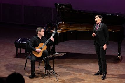 Nikola Printz, mezzo-soprano, in a masculine suit, in front of a piano, with Jon Mendle on guitar.