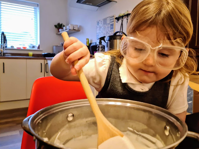 Image of a young girl in a school uniform wearing safety goggles over her eyes. She is sat on a red eames style chair and is mixing a white liquid (plaster of paris) inside a large metal stock pot with a wooden spoon.