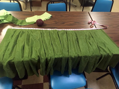 A pleated panel of green silk, draped over the edge of a brown plasticky table, with a pink measuring tape stretched above it, along with a red pincushion and a variety of green skirt pieces.