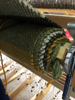 The cloth beam of a loom, with several layers of light green, marigold, dark green, and white striped fabric rolled onto it. The fabric continues diagonally up and to the left out of frame.