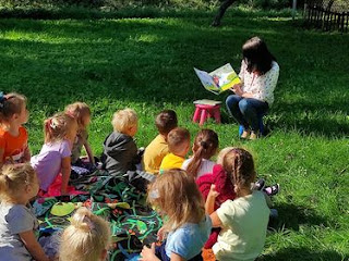 Grupa dzieci siedzi na kocu, który rozłożony jest na trawie. Dzieci słuchają pani bibliotekarki, która czyta im bajkę.