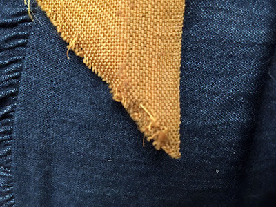 A close-up of a 45-degree point of mustard-brown fabric, with a selvedge on the right, vertical side and a frayed cut edge at left.