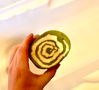 A dark green and pink swirled oval shaped bubble bar held by a white hand over a white oval bath tub on a bright background