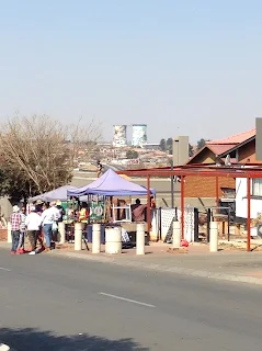 Market stall on the side of the road in front of a small home with a line of 10-12 people waiting to be served in front of it