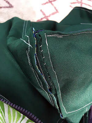 A close-up of the corner of a dark hunter green bag with white stitching, showing about two inches of hand sewing to hold the side and back edges together. The stitching lines mostly follow the edges of the pieces, but they're a little wobbly, and the backstitching only rarely aligns with the main line.