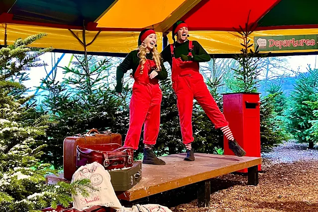 Two elves dancing on a small stage surrounded by Christmas trees