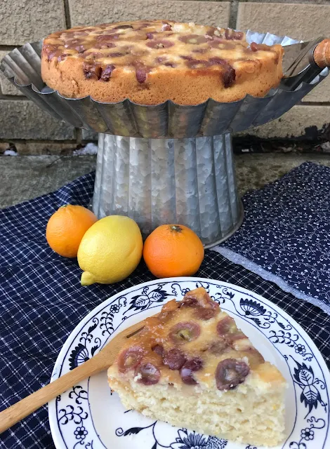 Side view of a cake stand with a finished grape upside down cake and a slice of finished cale on a small plate in front.