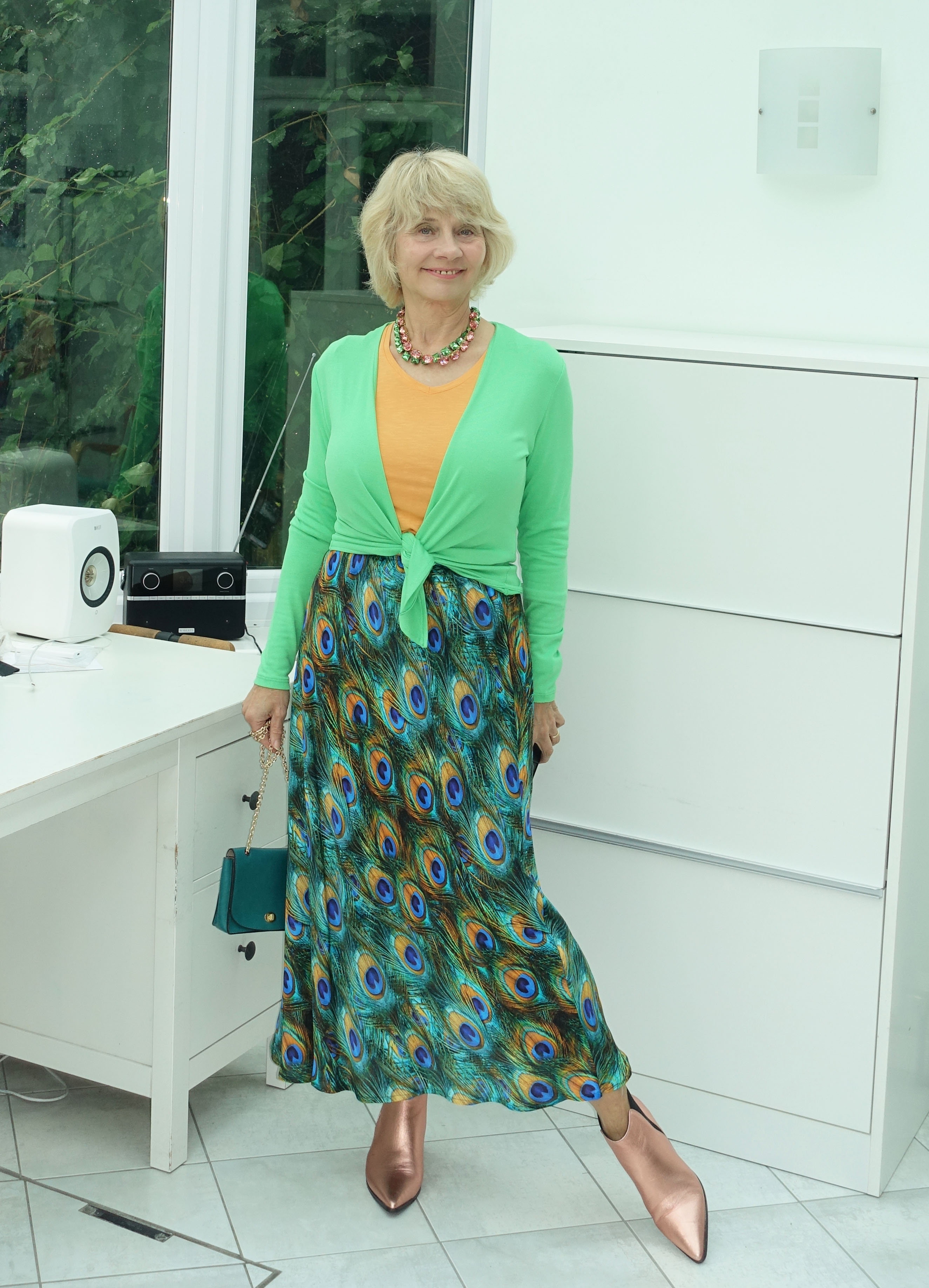 Gail Hanlon from Is This Mutton in peacock patterned skirt, green short tie wrap, bright apricot top and rose gold ankle boots.