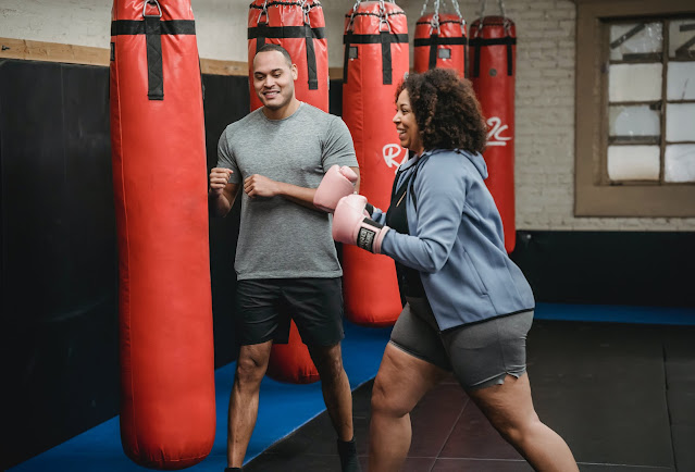 A cheerful ethnic trainer is seen training with a fit and laughing Black woman in a gym, with weights and exercise equipment in the background.