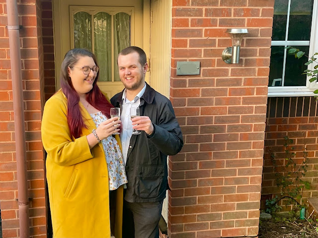 Becky and Jack are holding glasses of prosecco and clinking glasses outside their new home