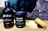 A wide shot photo of a tall cylindrical bottle filled with dark green liquid with a black label that says grass shower gel lush in white font on a bright background next to a black wide cylidericnal pot that says karma cream lush in white font filled with orange cream next to a yellow crown shaped solid body butter and a purple circular shampoo bar with yellow flowers on  top on a bright background