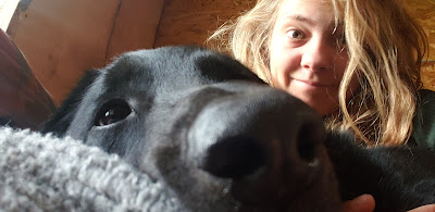 Image shows a selfie of Lisa and Monty. It is a very close up of Monty's nose/snout as he lies on the couch cuddling with Lisa. He looks tired/pensive in the photo, and Lisa is making a smirky face behind him, enjoying the couch cuddles!