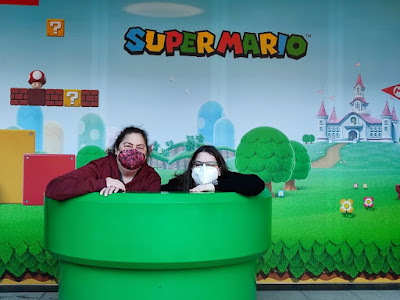 Grüne Röhre, darin Miriam und Annabell, beide tragen FFP2 Masken. Hintergrund ein Bild aus dem Super Mario ComputerSpiel mit bunten Röhren, Bäumen, einem Schloß, Steinblöcken, eine Fahne, Blumen, ein Pilz und der Schriftzug "Super Mario".