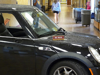 A black car is parked in a building. A desk name plate (which reads "Mr. Oblas") rests on the hood of the car.