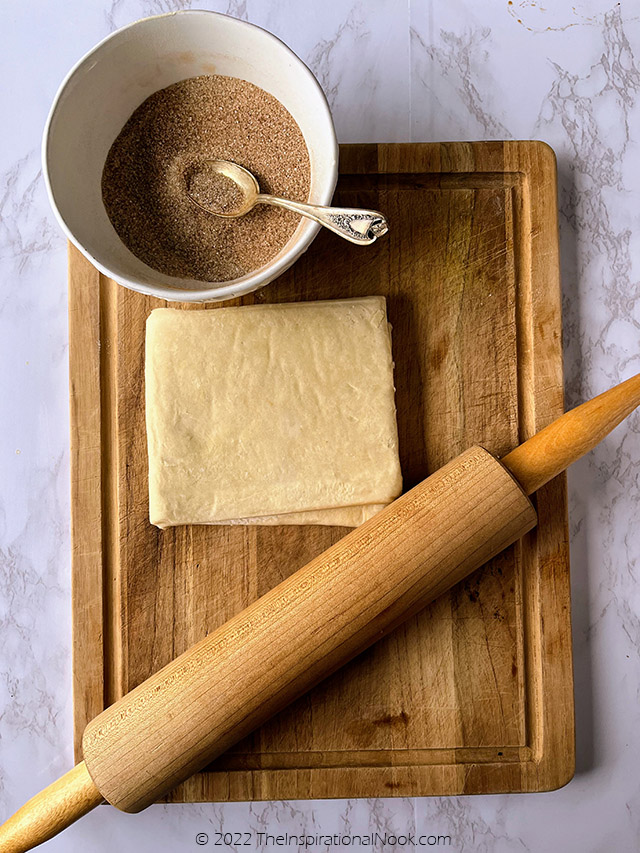 Rough puff pastry on a wooden board with a rolling pin and a white bowl filled with cinnamon sugar