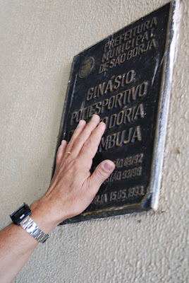 Uma placa com o nome do “Ginásio Cleto Dória de Azambuja” com letras em relevo. A foto enquadra apenas a placa e a mão de Ramão em cima das letras. Ele usa um relógio no pulso esquerdo.