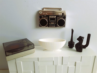 One-twelfth scale modern miniature sideboard with a turntable, large planter/vase ans selection of mid-century ornaments on it. On the wall above is a decorative piece in the shape of an 80s boom box.