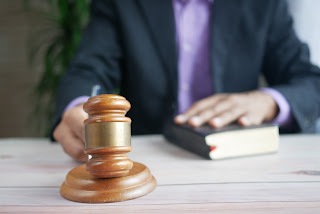 Image from a court room where a males shirt and jacket can be seen, as well as his hand holding a gavel and a book placed under his other hand.