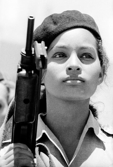 Uma garota é vista em posição militar, vestindo camisa e boina segurando um rifle com a mão esquerda. Seu olhar está voltado para o lado direito da imagem e sua expressão é séria.