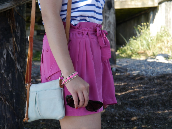 A more detailed view of a striped tee worn with fuchsia silk shorts, bracelet, and a small but colourful crossbody bag and cat-eye shades.